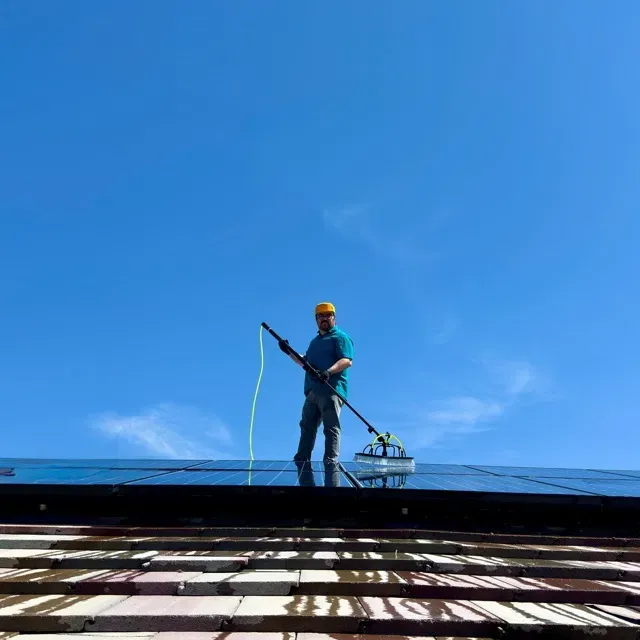 Technician cleaning a residential solar array with a water-fed pole under a clear blue sky.